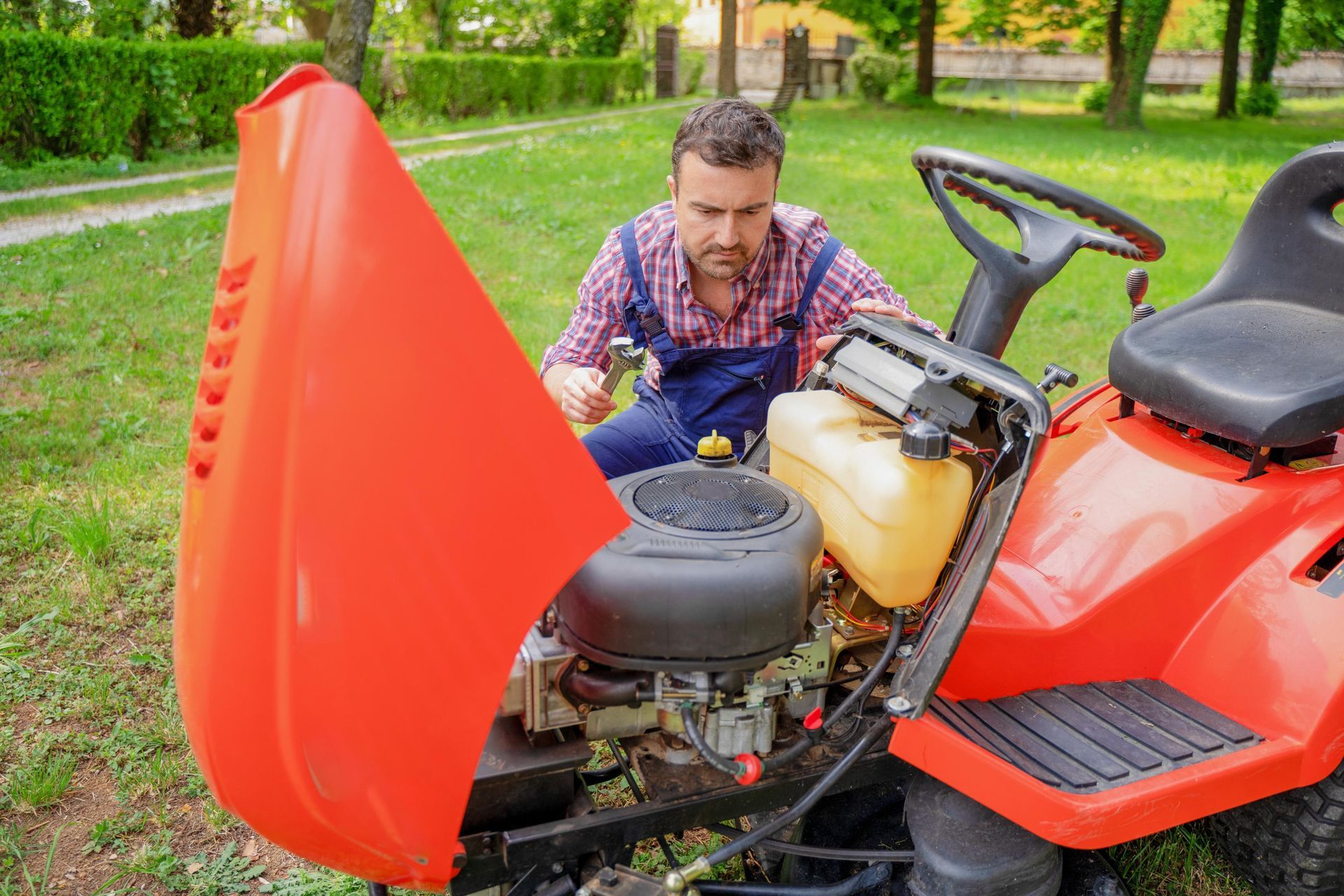 Man repairing a red ride-on mower with its hood open in a grassy yard