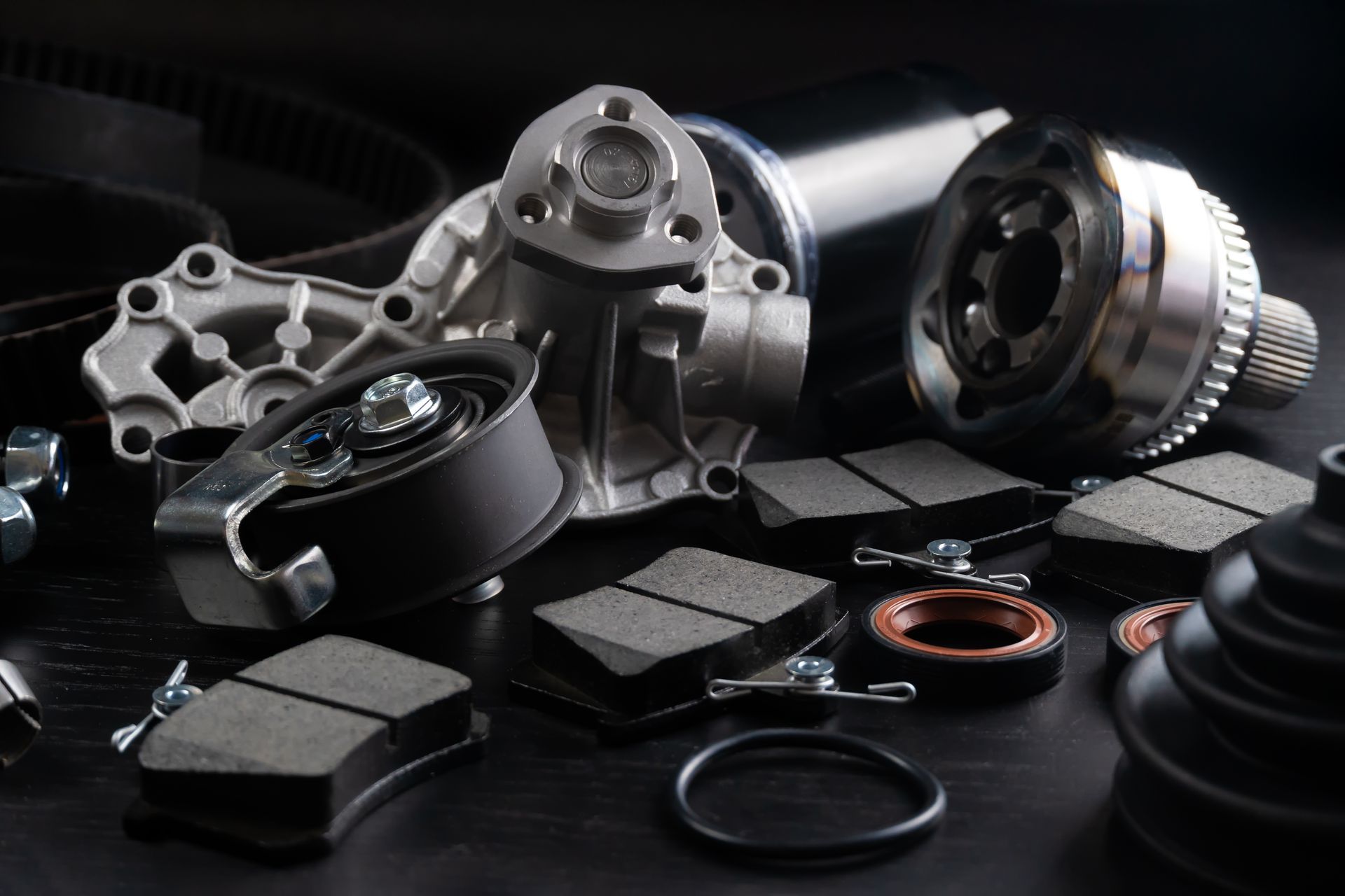 Assorted automotive parts, including brake pads, pulleys, seals, and a water pump, on a dark surface