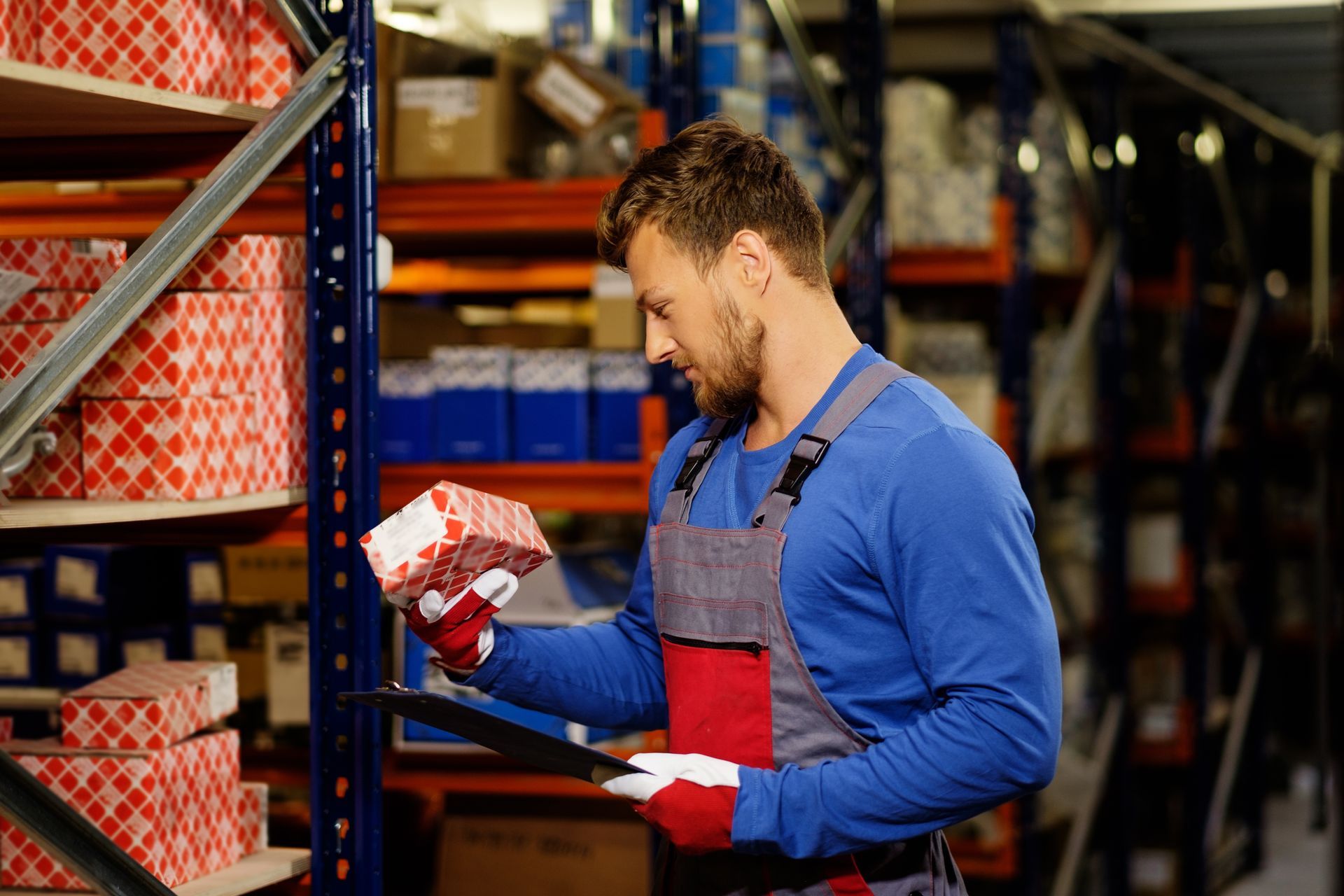 Warehouse worker checking a boxed item against inventory on shelves with a clipboard.
