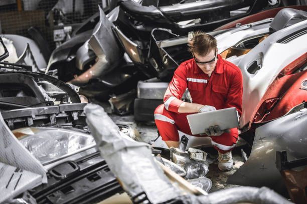 Auto technician inspecting damaged car parts in a scrapyard using a tablet for assessment.