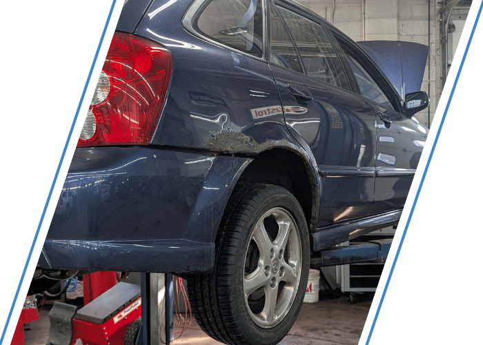 Blue wagon lifted for automotive repair with visible body rust near the rear wheel.