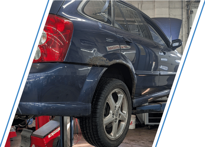 Blue wagon lifted for automotive repair with visible body rust near the rear wheel.