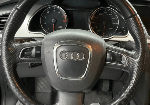 Close-up of an Audi steering wheel and instrument cluster inside the vehicle, highlighting the driver's controls and dashboard gauges.