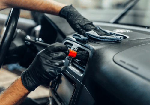Technician cleaning a vehicle's dashboard and air vents with a detailing brush and microfiber cloth