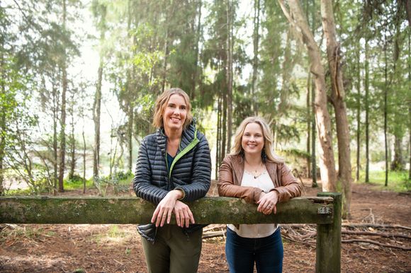 Vanessa and Lana leaning on a wooden fence in a sunlit forest clearing, smiling at the camera.