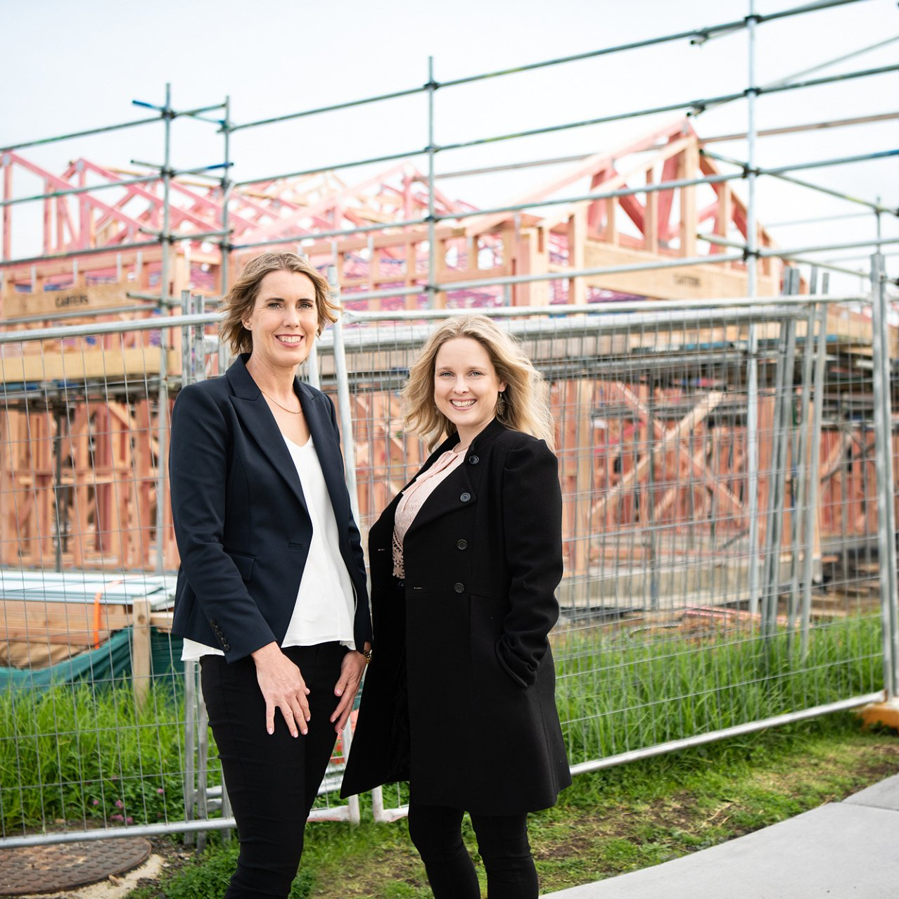 Vanessa and Lana standing in front of a fenced construction site with building framework and scaffolding behind them