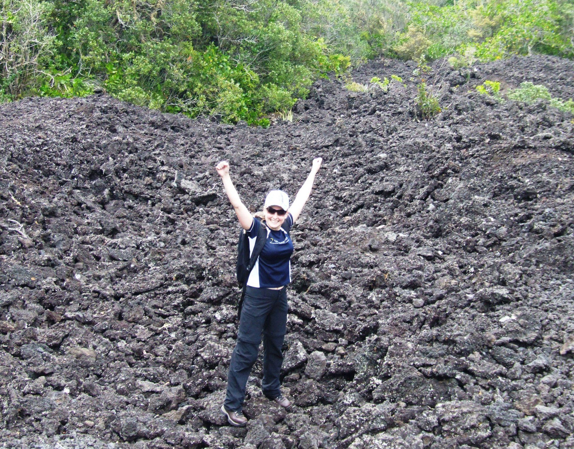 Lana with arms raised standing on dark rocky terrain with green foliage in the background