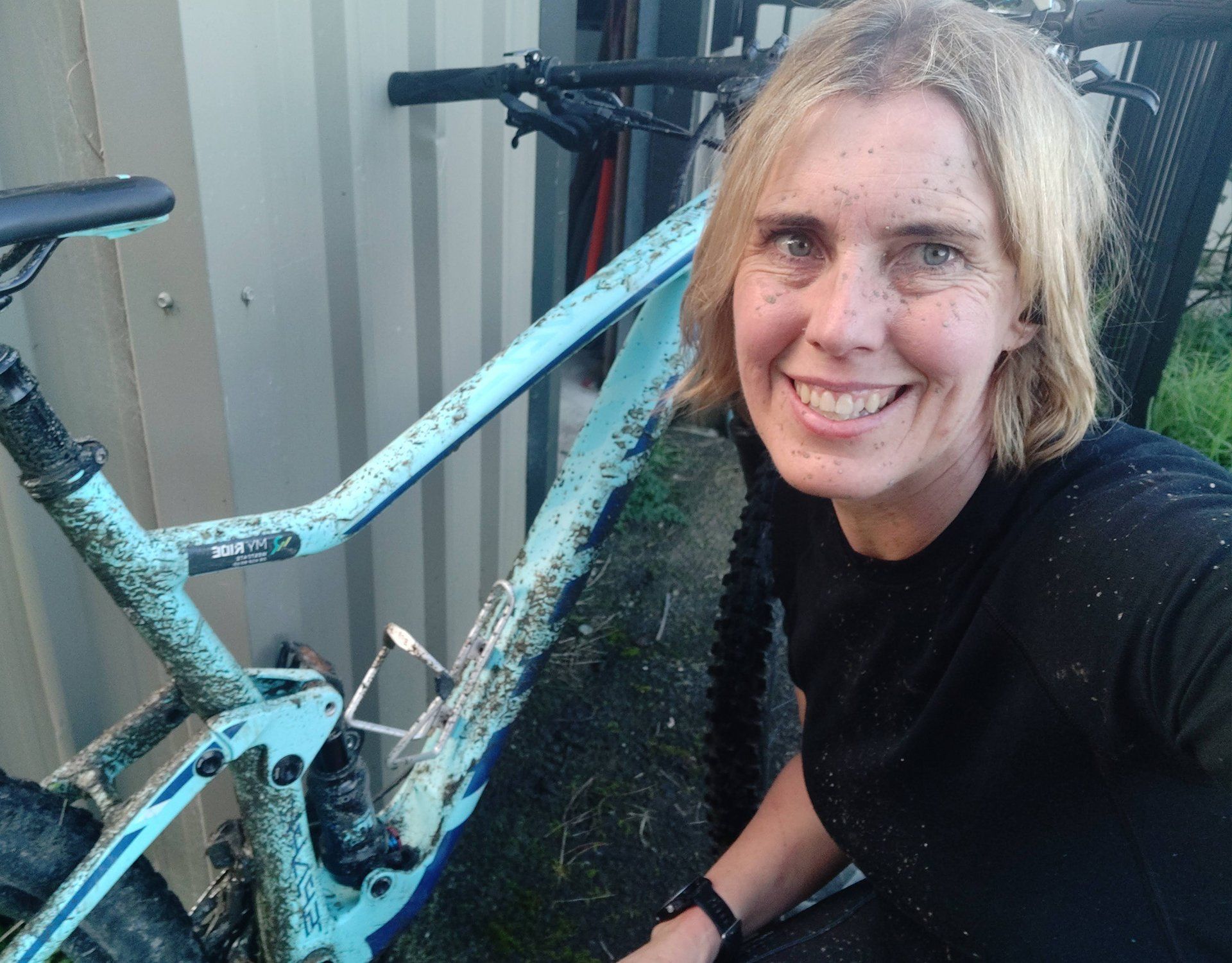 Vanessa - Smiling person beside a muddy blue bicycle outdoors, with a shed and other bikes in the background