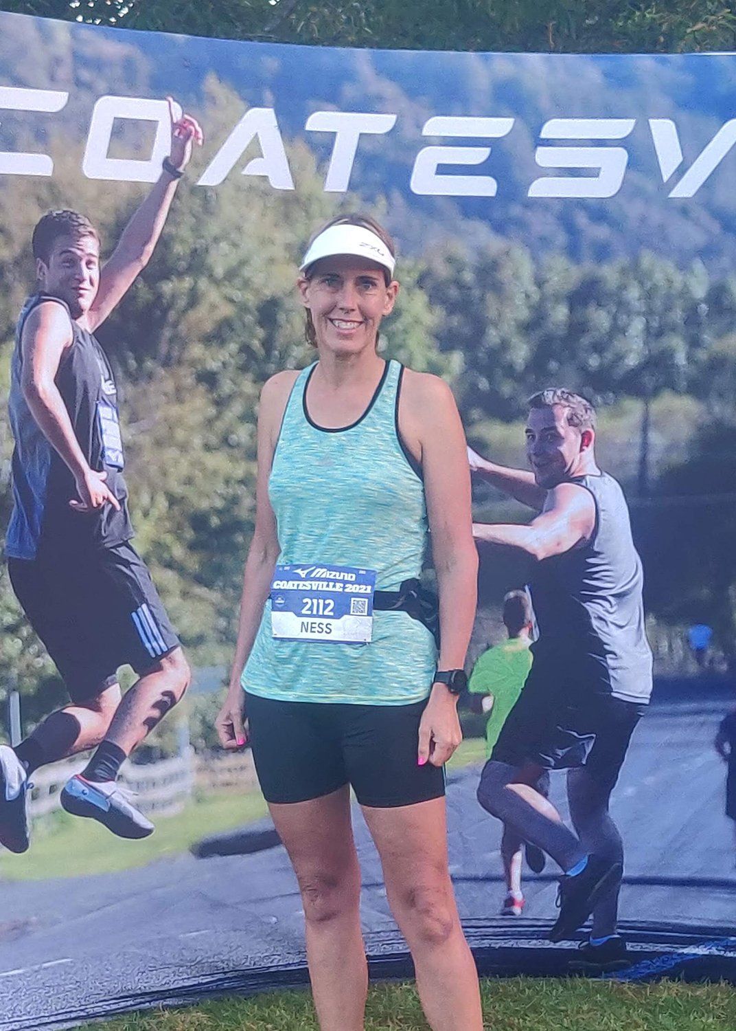 Vanessa - Woman in green tank top and cap posing at a finish line banner outdoors, with runners in the background