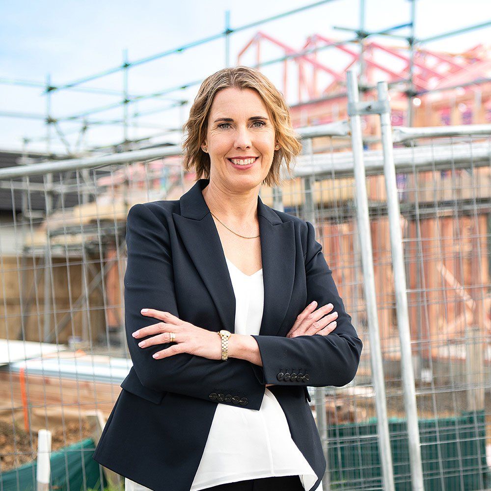 Vanessa standing with arms crossed at a construction site, wearing a navy blazer and smiling.