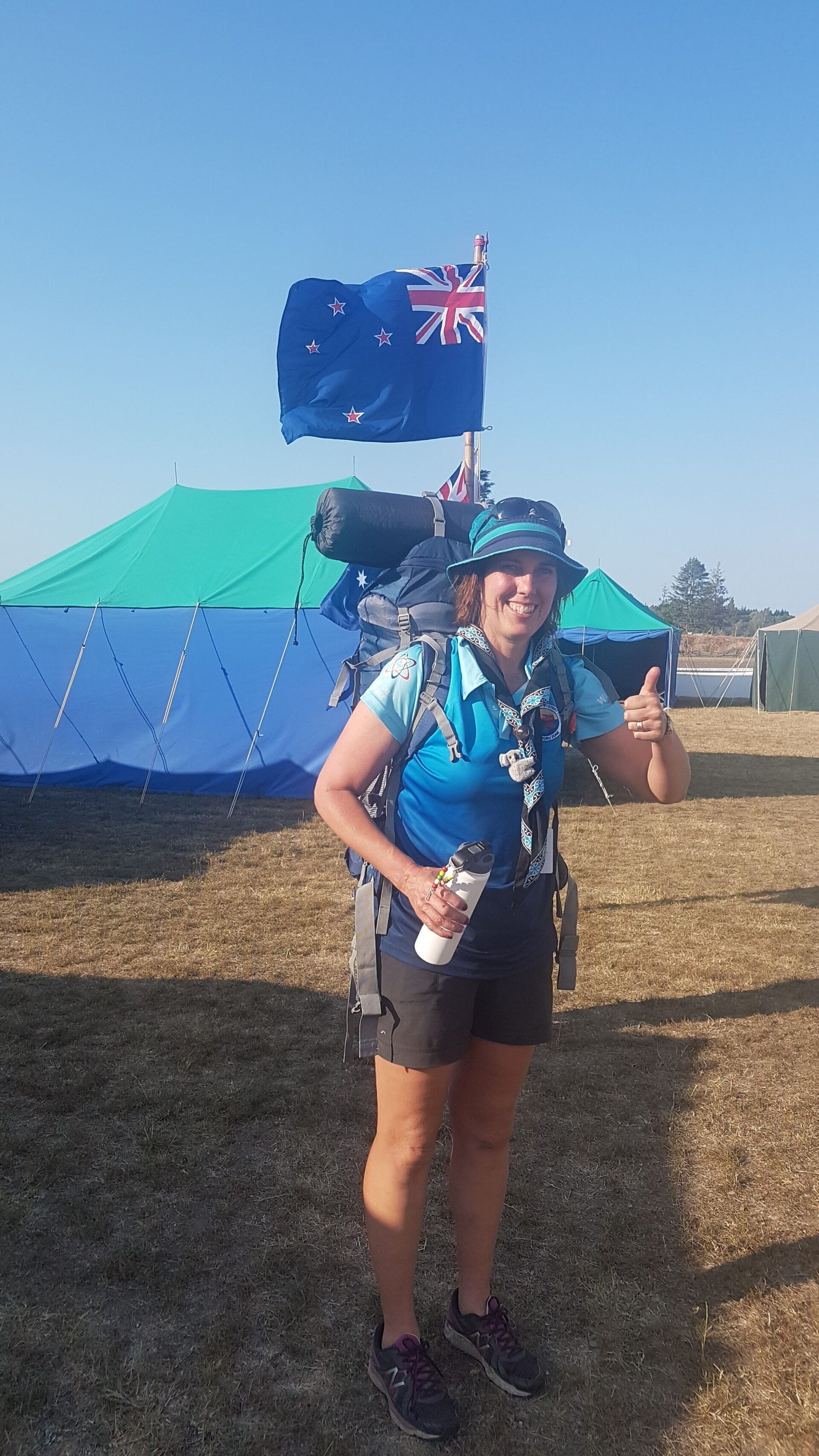Person in blue hiking gear stands before a tent, holding a flag in a sunny outdoor campsite.