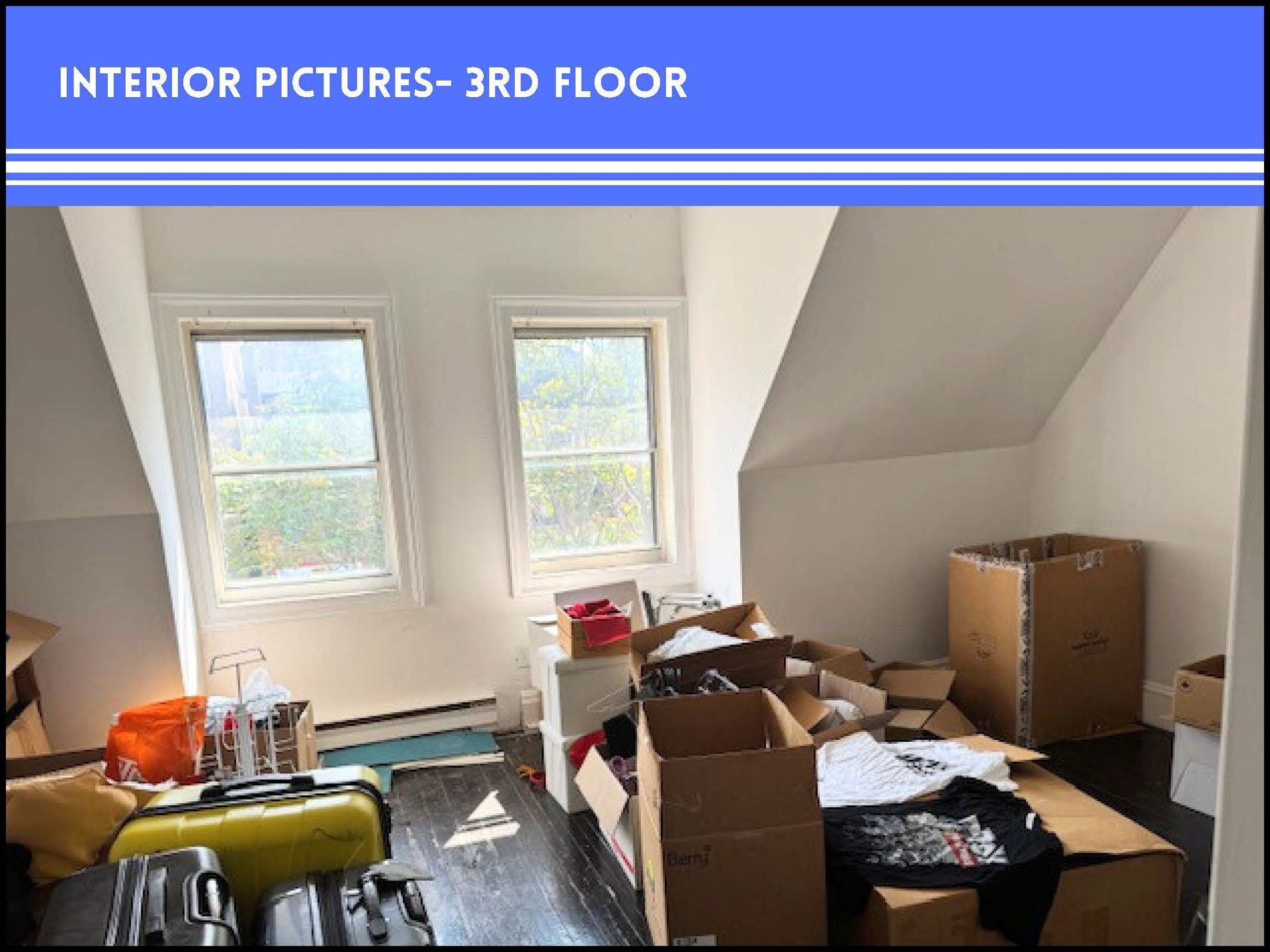 A third-floor room with sloped ceilings, two windows, and several cardboard moving boxes and suitcases on the floor.