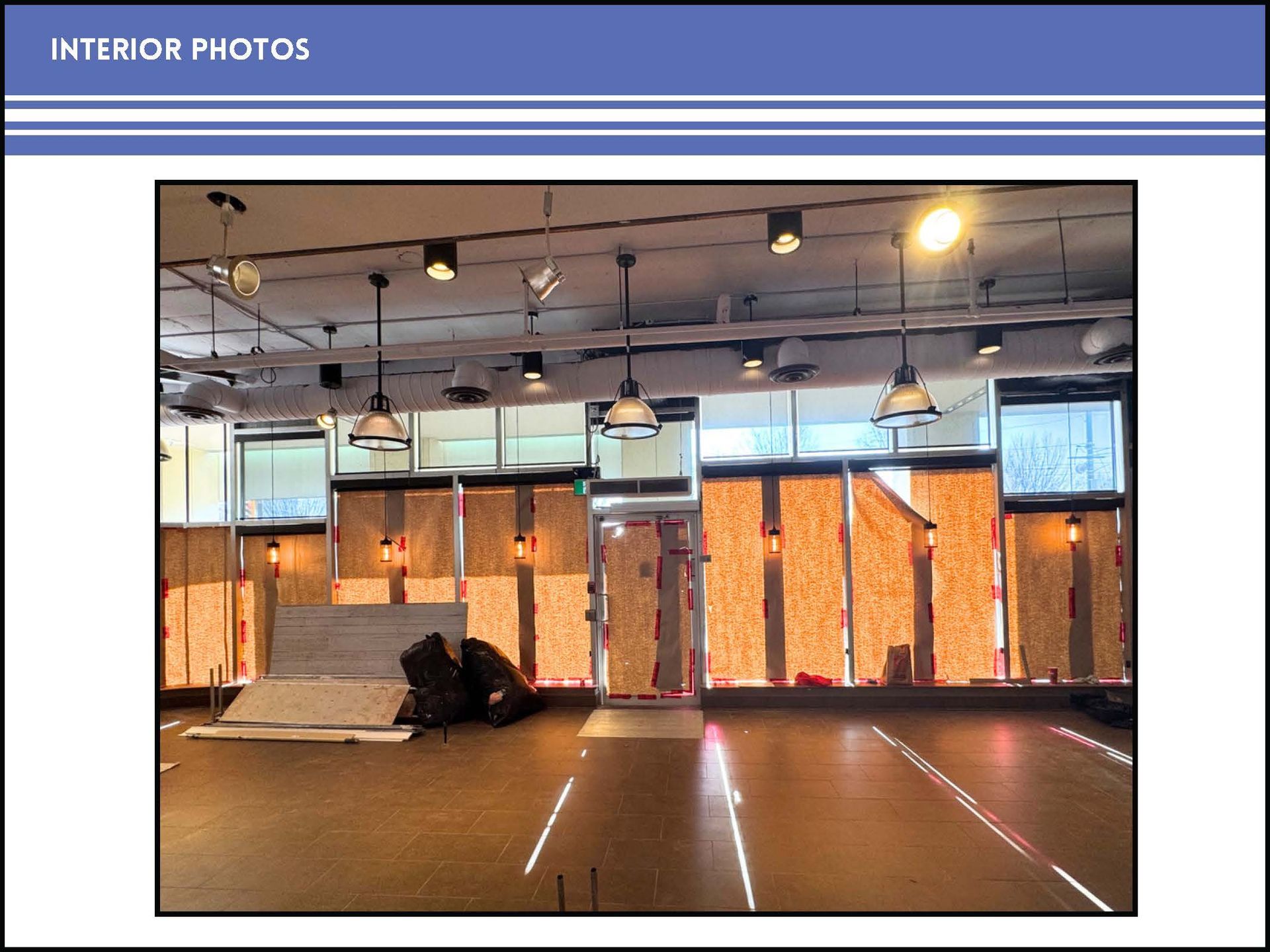 An interior view of a vacant commercial space with large windows boarded up with plywood and hanging pendant lights.