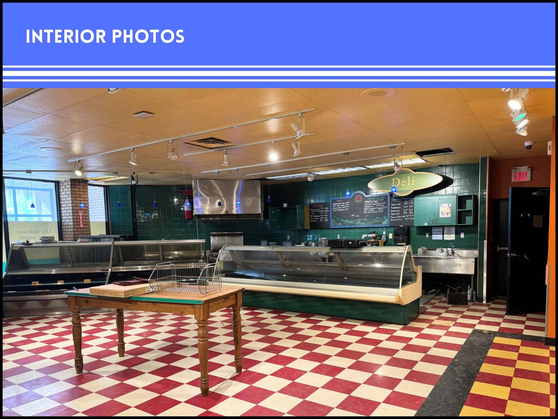 An empty deli interior with a checkerboard floor, a wooden table, display counters, and a green wall with signage.