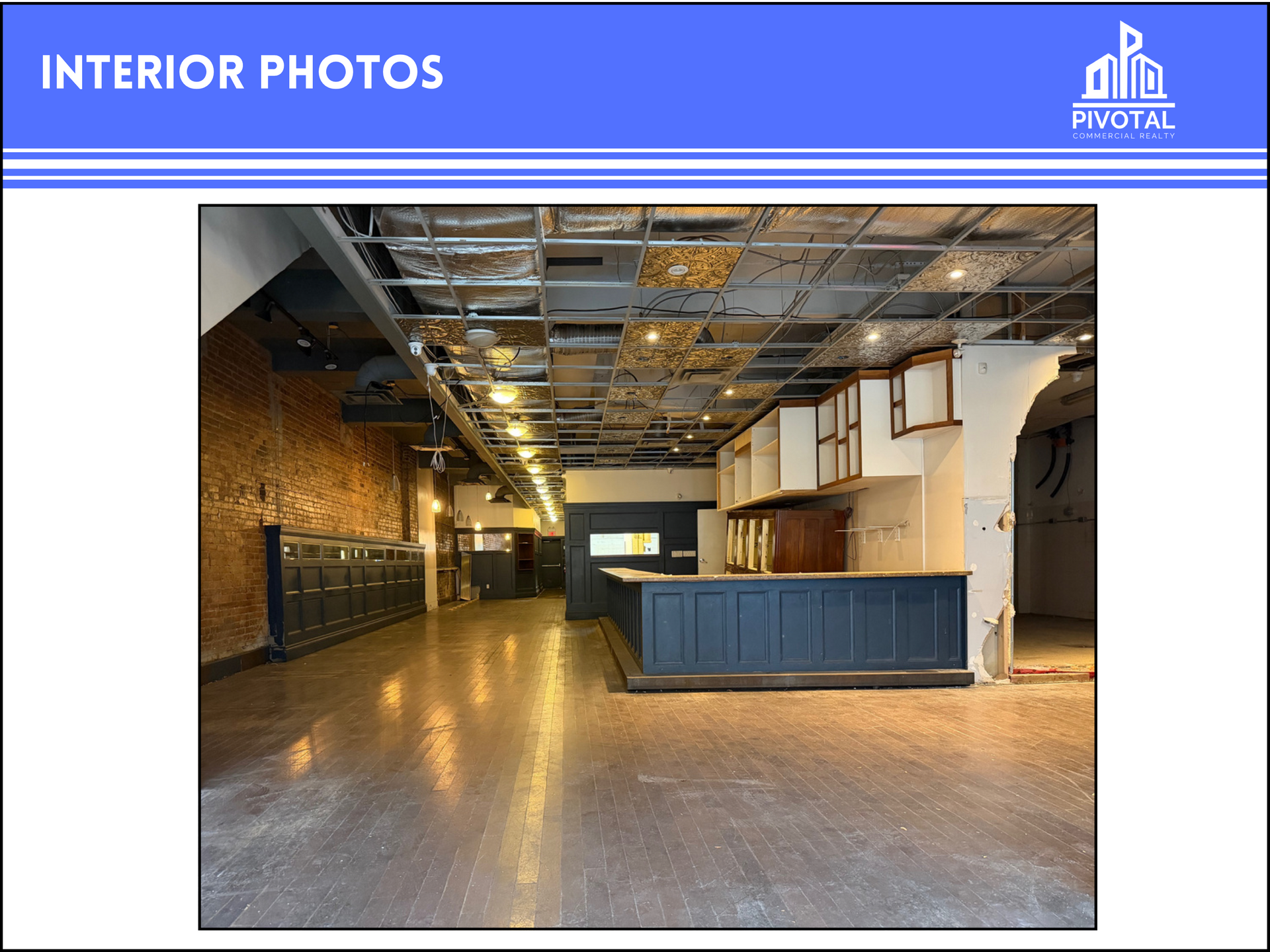 An interior view of a vacant commercial space with exposed ceiling grid, brick wall, and a dark bar counter.