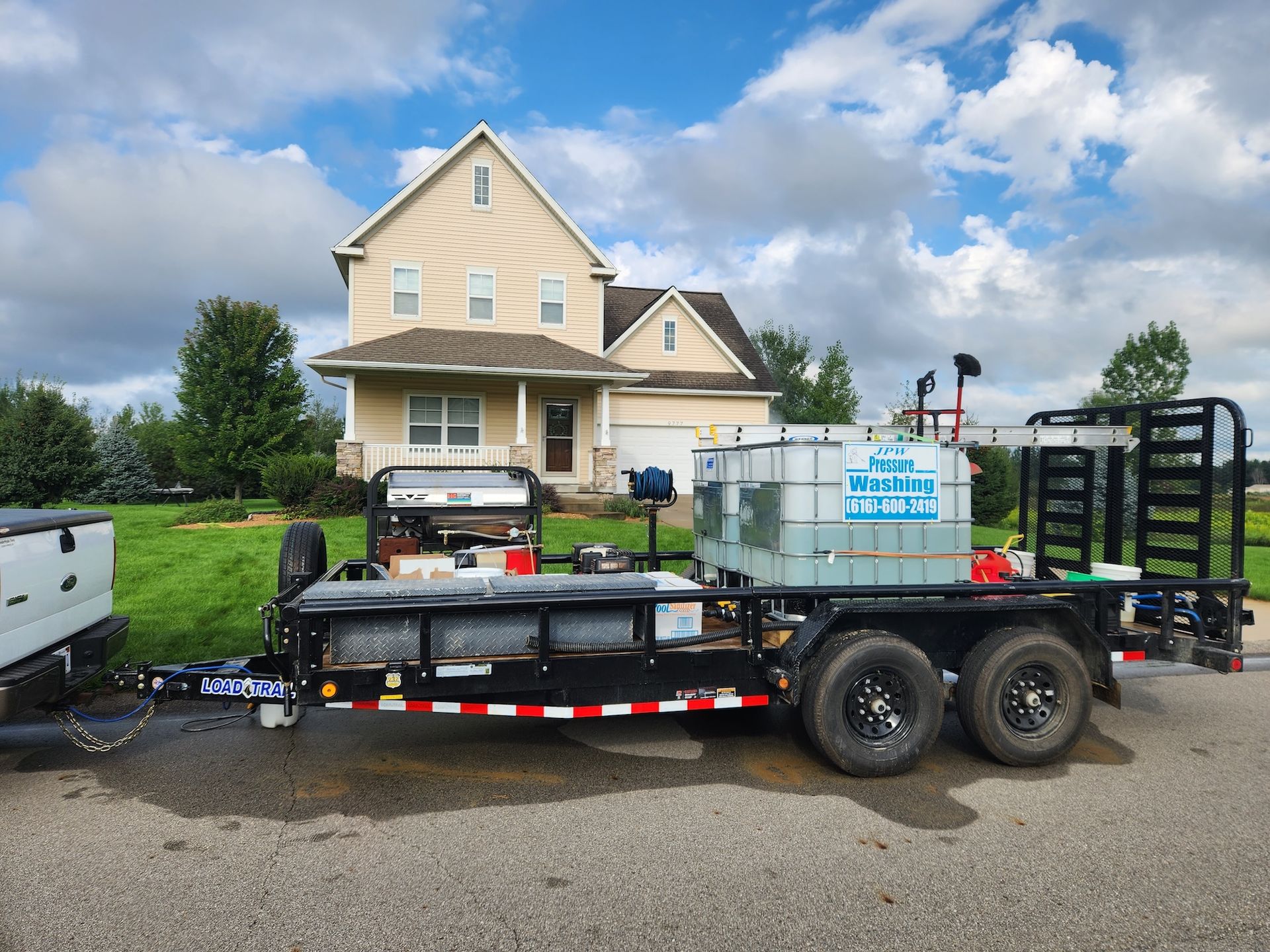 A trailer with equipment, parked in front of a beige house with a cloudy sky overhead.