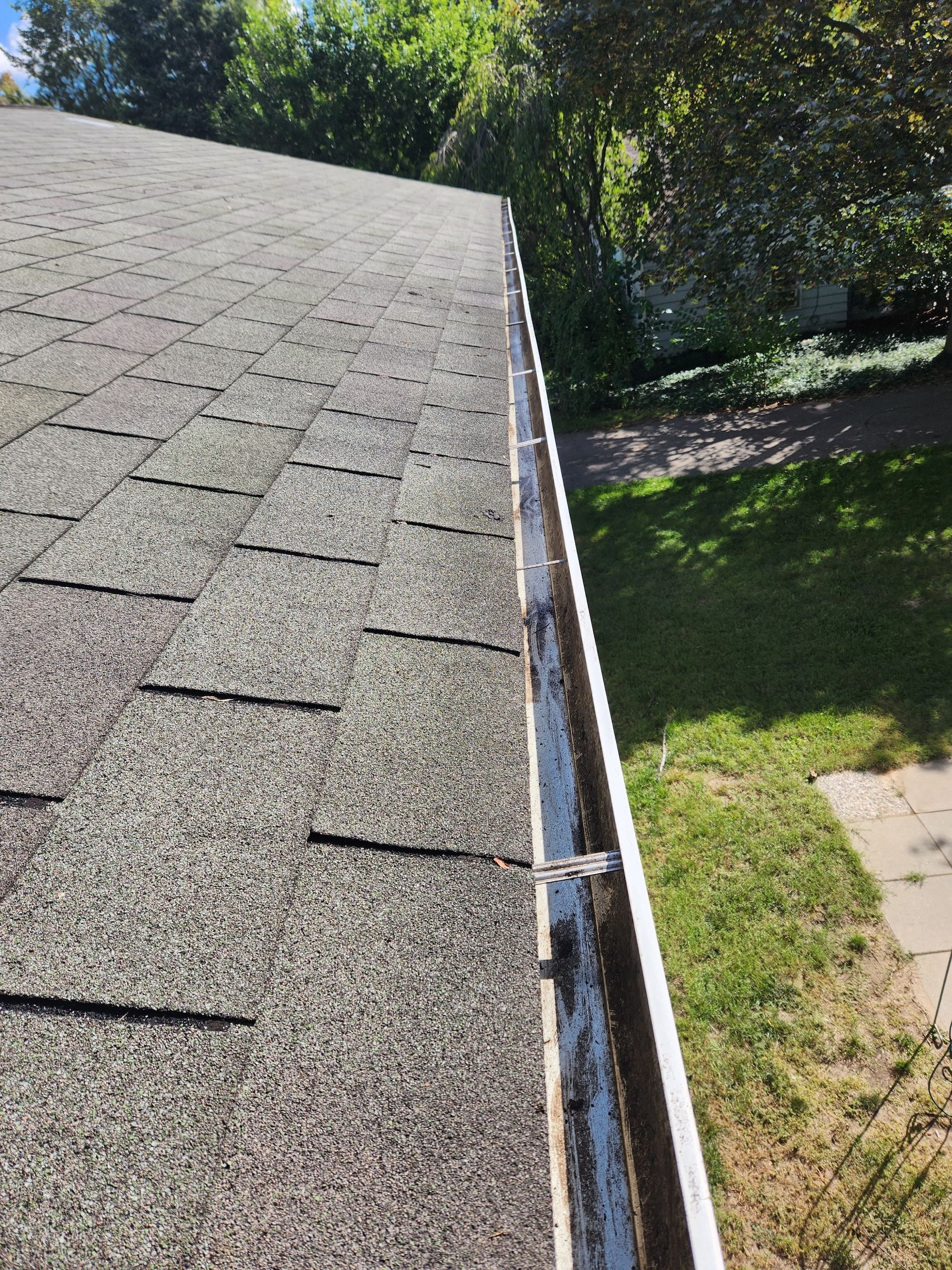Roof with shingles and gutter filled with debris, with a grassy area in the background.