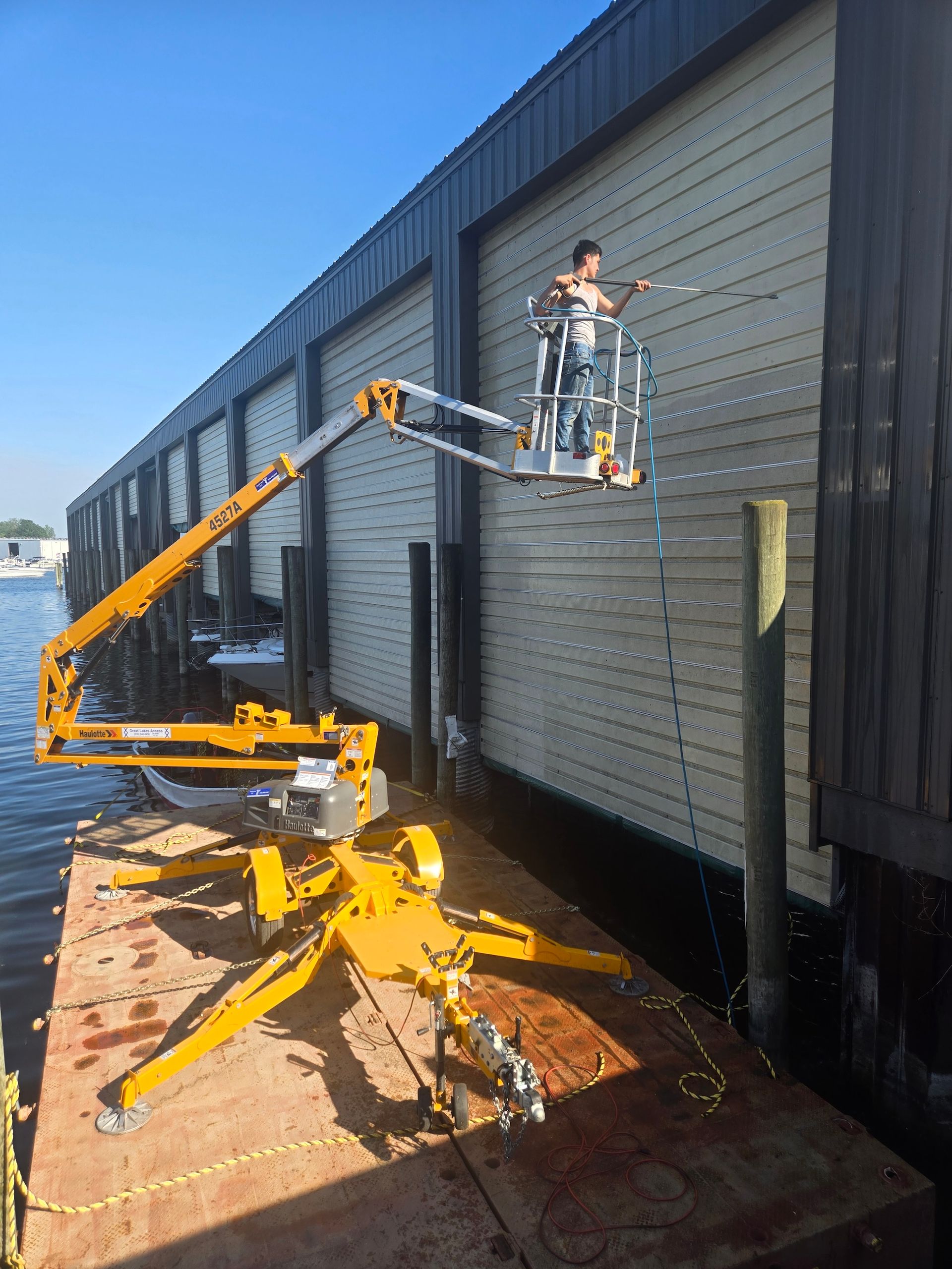 Yellow lift platform on a dock, person in the basket spraying a building. Sunny day.