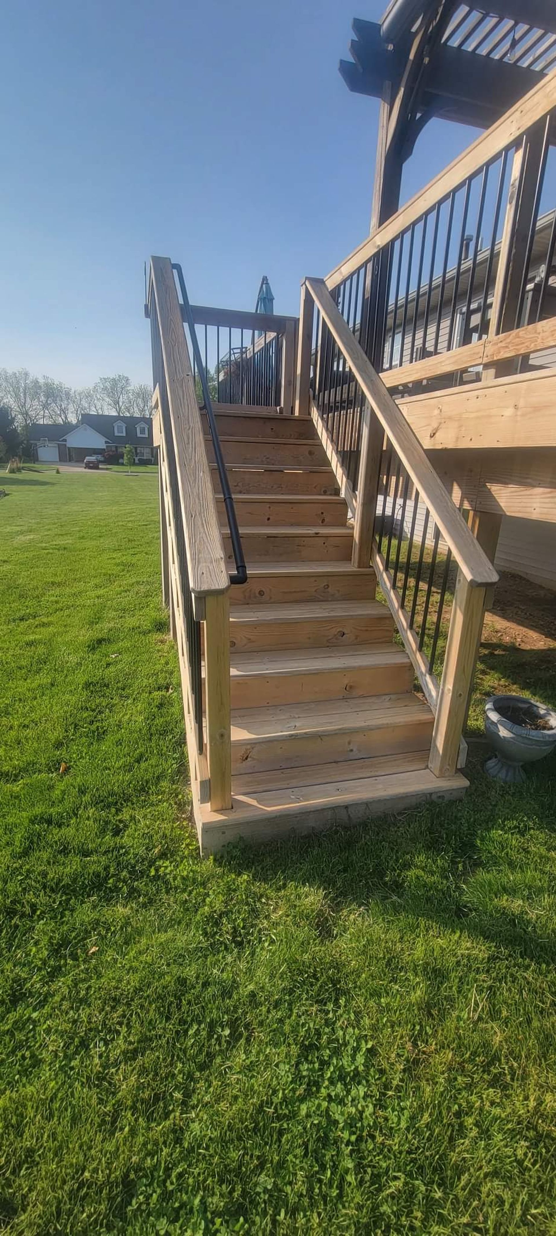 Wooden outdoor staircase leading up to a deck, set in a grassy yard under a bright blue sky.