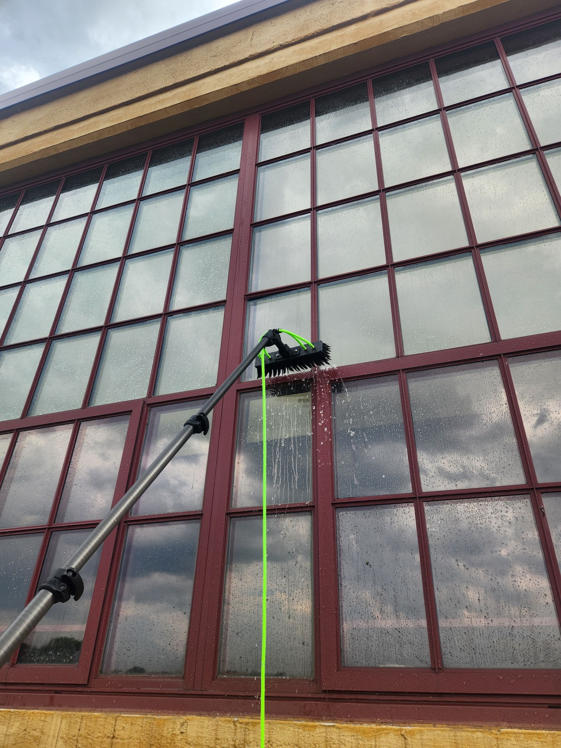 Cleaning a large window with a long-handled brush. Dark red window frames, soapy water, and cloudy sky visible.