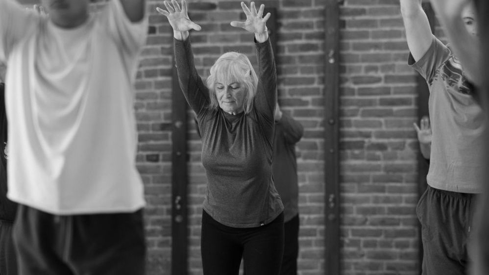 People in a fitness class. An older person stretches arms up, others blurred in the background, brick wall.