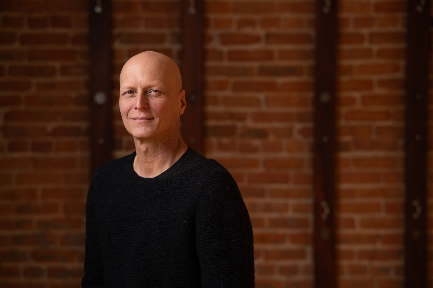 Man with shaved head in black sweater smiles in front of a brick wall.
