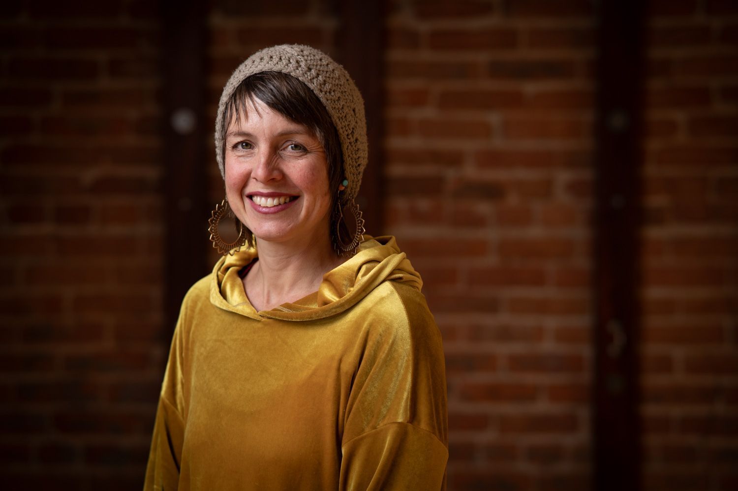 Woman in mustard yellow hoodie and knitted hat smiling in front of a brick wall.