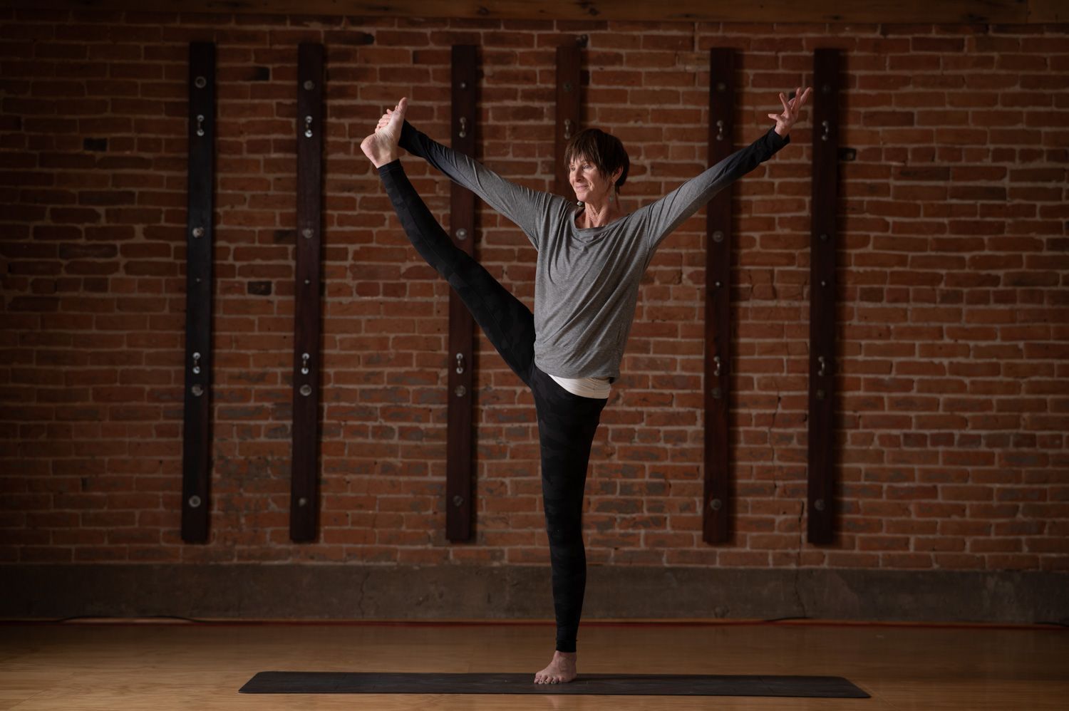 Woman balances in yoga pose, standing with leg extended up, arms outstretched. Brick wall background.
