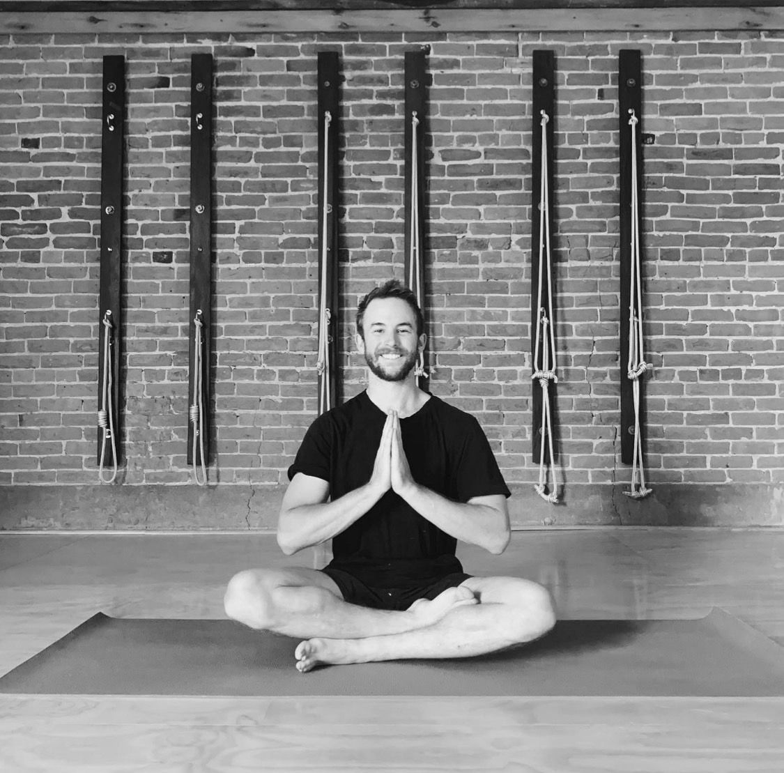 Man in lotus position with hands in prayer pose, in a studio with exposed brick and yoga equipment.