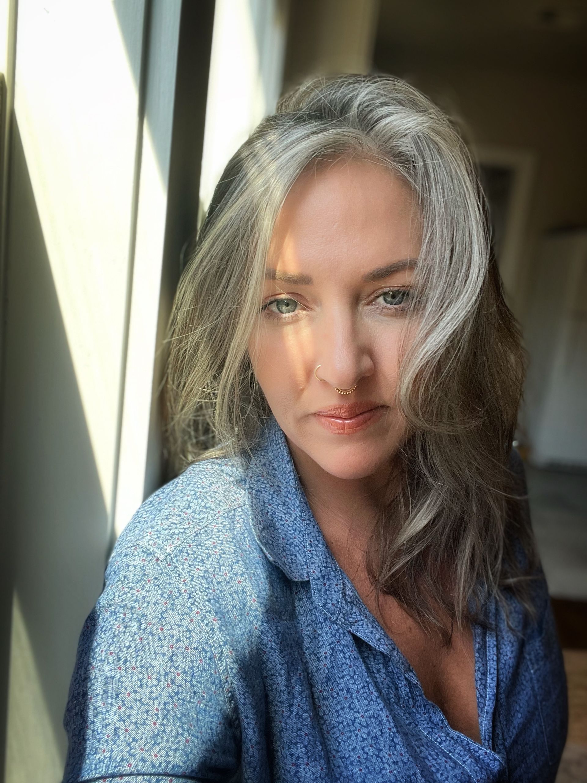 Woman with graying hair, blue shirt, gazing towards camera, lit by sunlight.