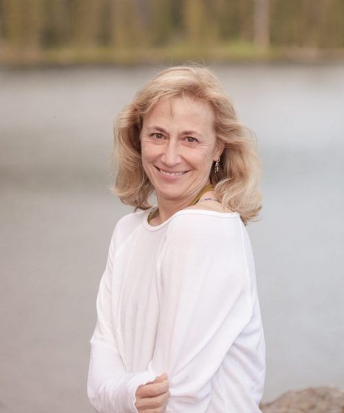 Woman with blonde hair smiles, wearing a white sweater, near a body of water, soft focus background.