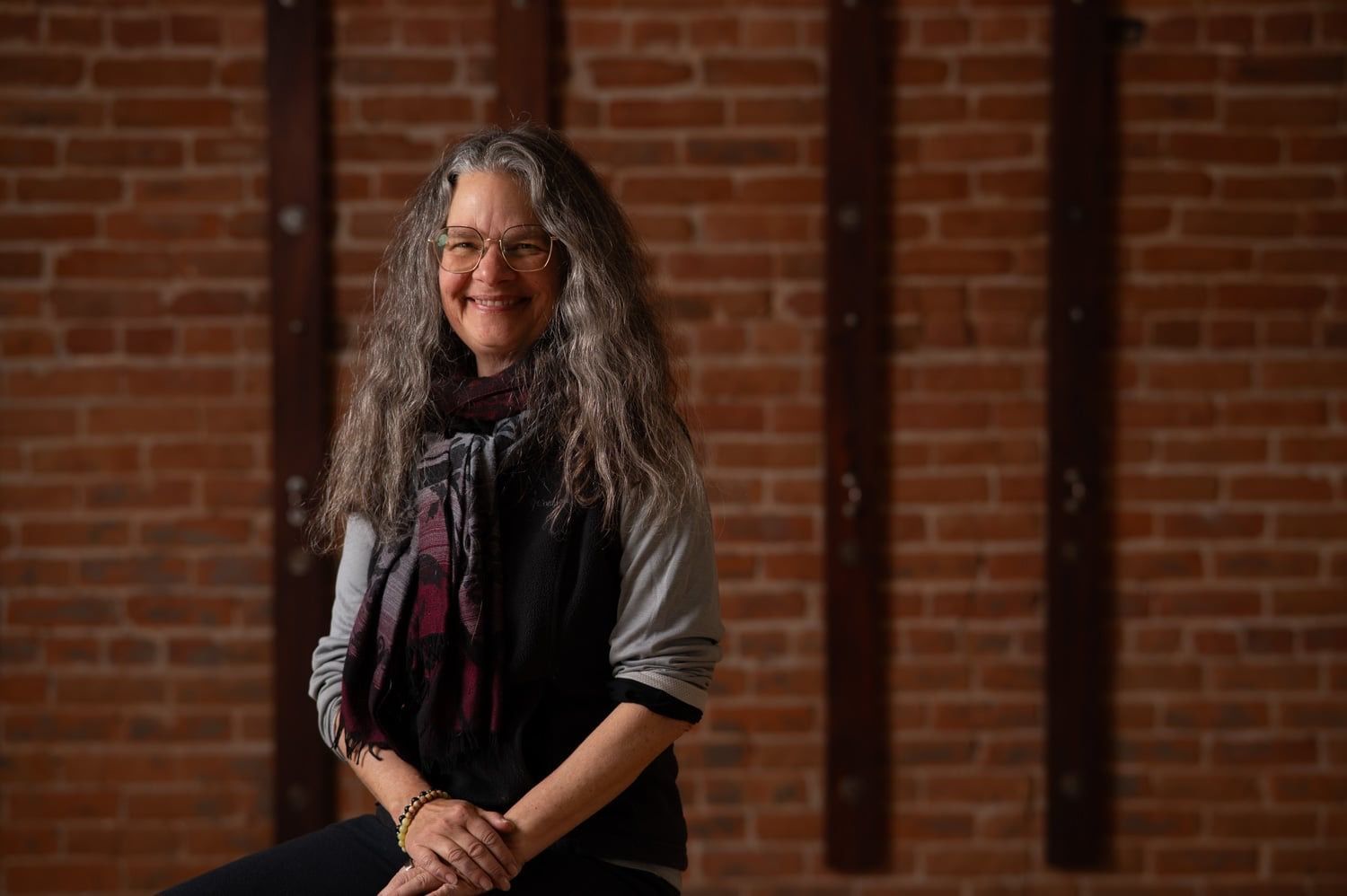 Woman with long gray hair smiles, seated in front of a brick wall.