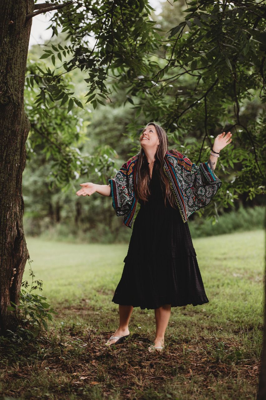 Woman in black dress and colorful shawl outdoors with arms outstretched, looking up at trees.
