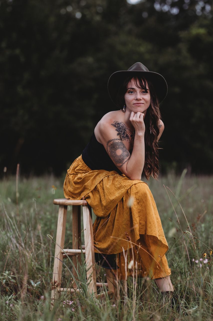 Woman wearing a hat and yellow pants sits on a stool in a field, smiling, arm tattoos visible.