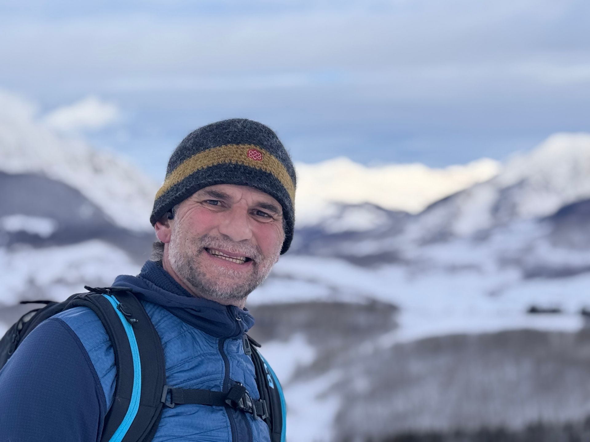 Man in blue vest and hat smiles in snowy mountains.