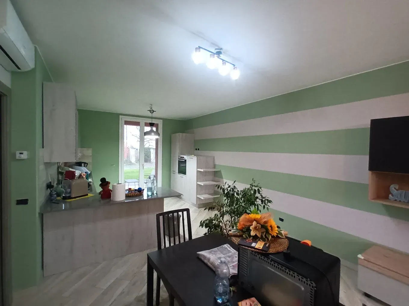 Cucina moderna con parete a strisce verdi, tavolo da pranzo e fiori vicino a una finestra.