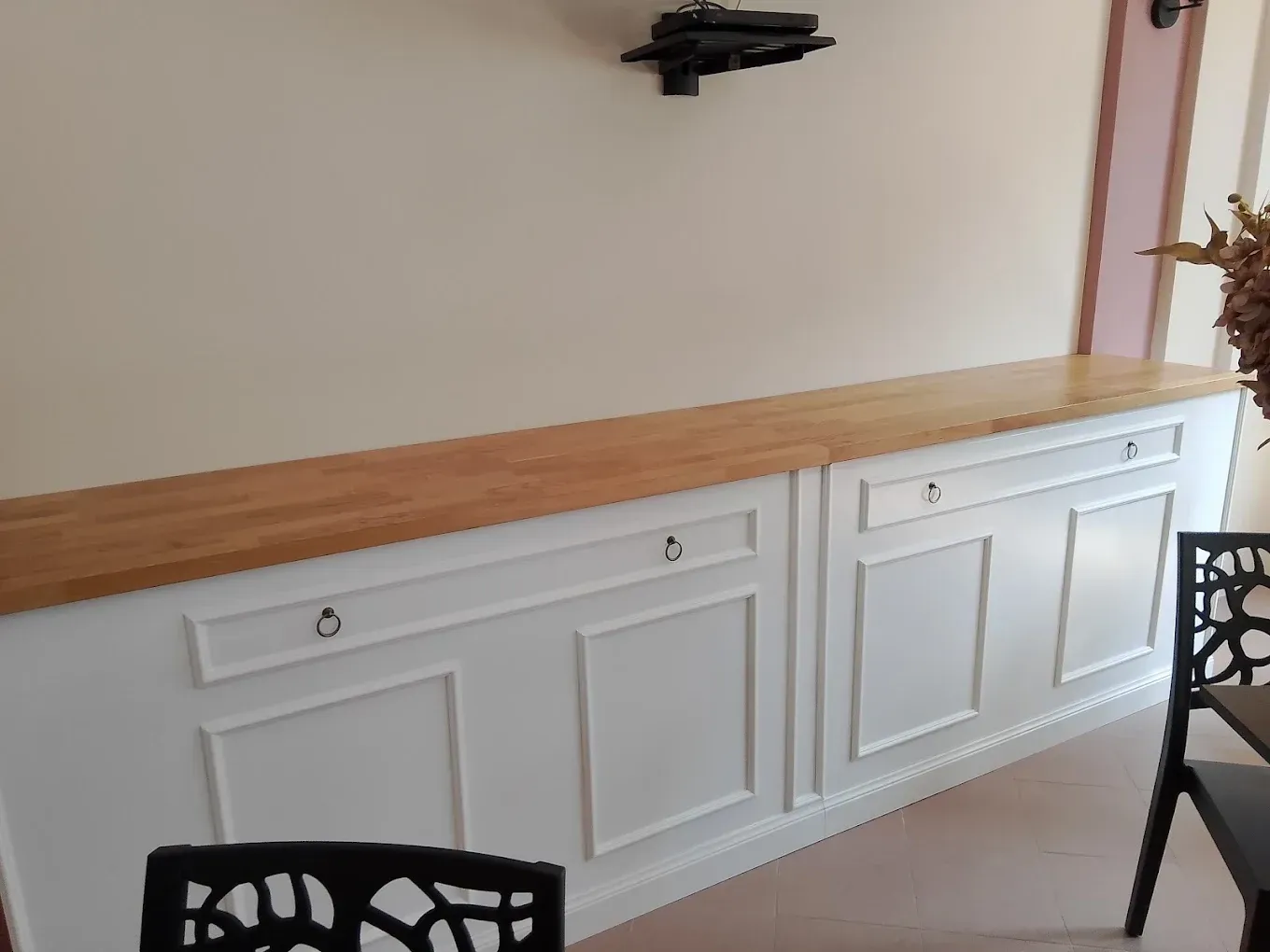 Mobile a muro bianco con piano in legno in una zona pranzo, con sedie nere parzialmente visibili.