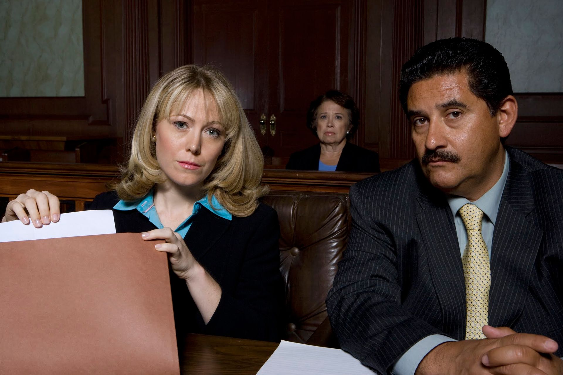 Two attorneys in courtroom: one blonde woman holding paper, one man with mustache looking serious. A judge in background.