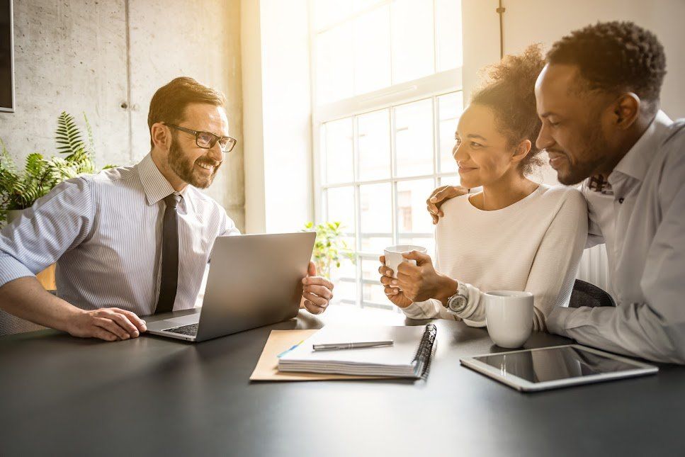 Financial advisor showing information on a laptop to a couple at a table; sunny room.