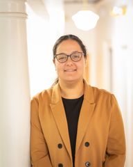 Monica W. Henain Woman with glasses, leaning against a white pillar, wearing a brown blazer, black shirt, smiling in an indoor hallway.