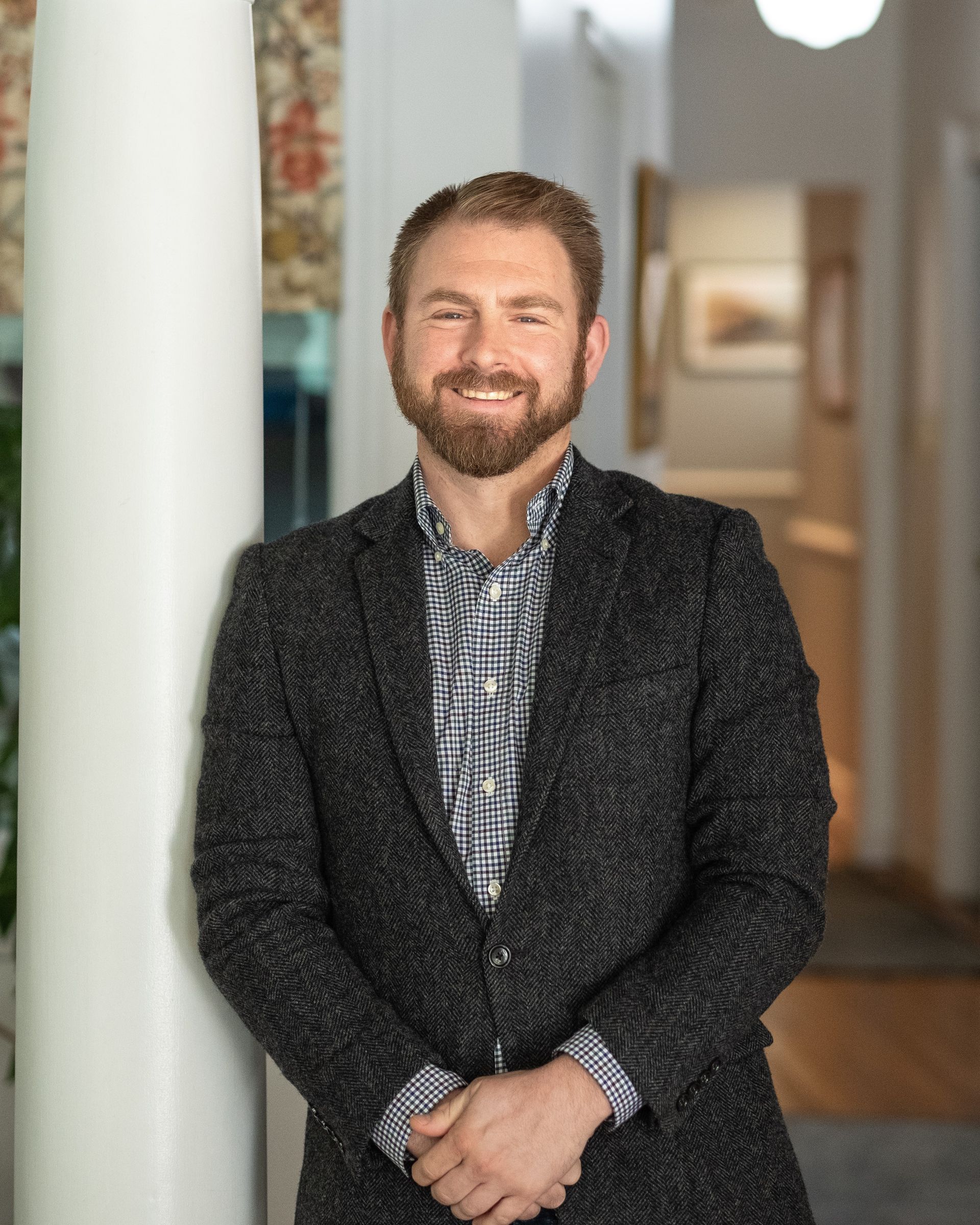 Man with beard, leaning against a white pillar, wearing a patterned shirt and dark jacket in a hallway.
