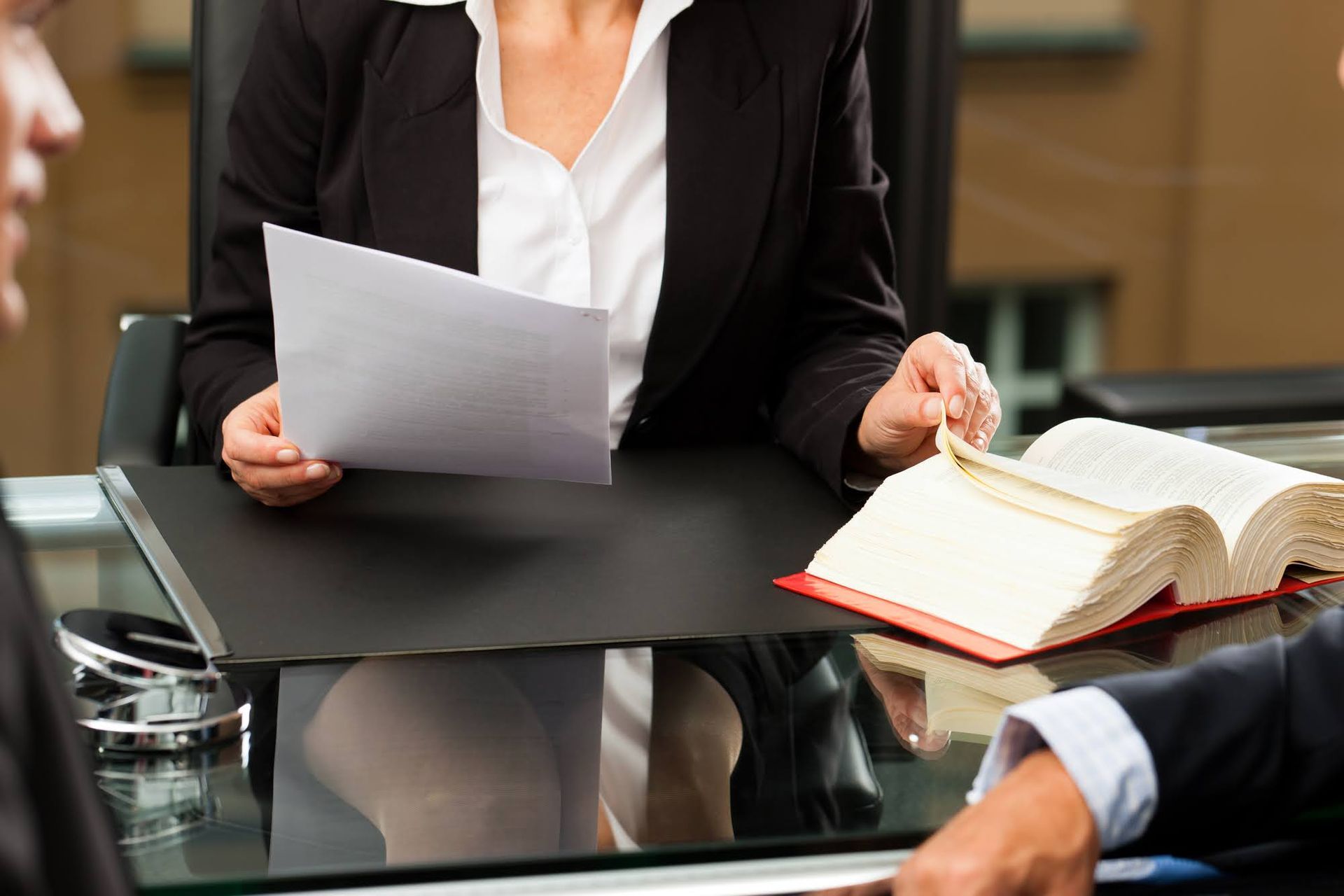 Woman in black suit reviewing documents, open law book on desk with others.