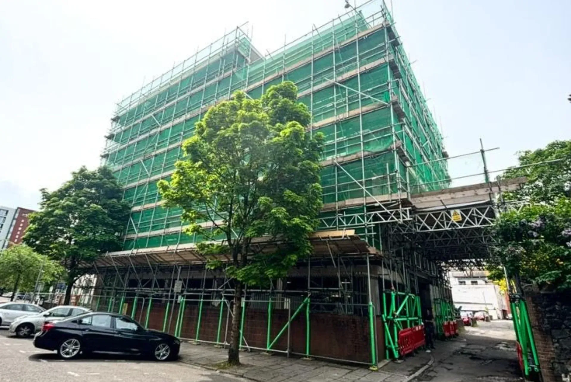 Green scaffolding around a building under construction beside parked cars and a tree on a street corner.