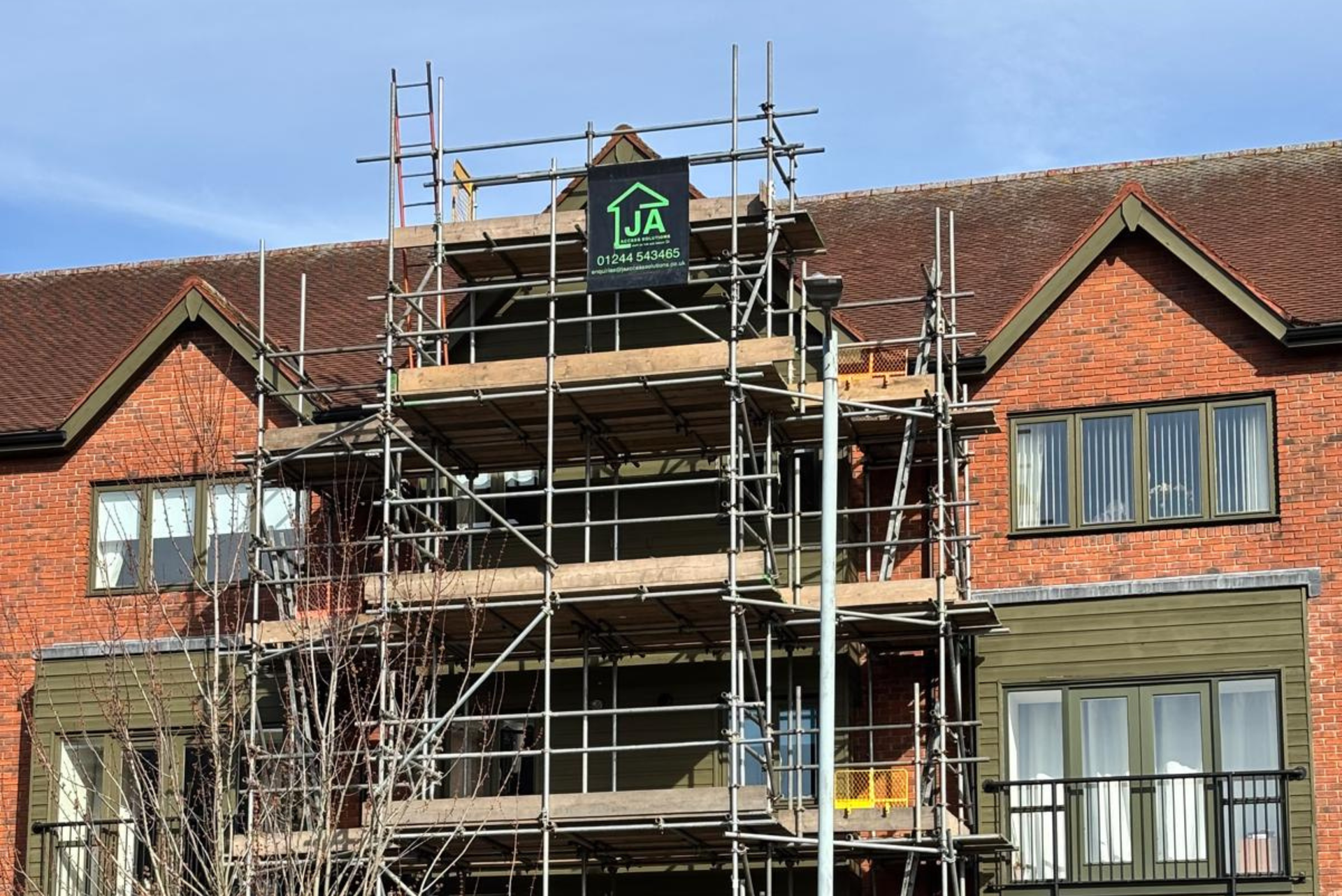 Brick house under construction with scaffolding and a sign on the roof against a blue sky