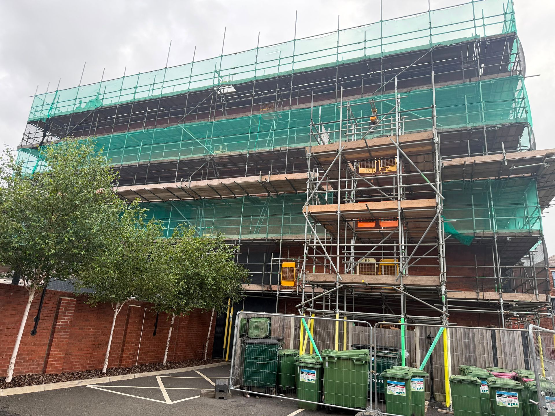 Green scaffolding around a building under construction beside parked cars and a tree on a street corner.