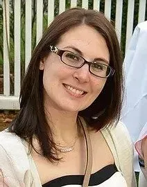 Woman with glasses smiles, tan cardigan, smiling, near a white fence.