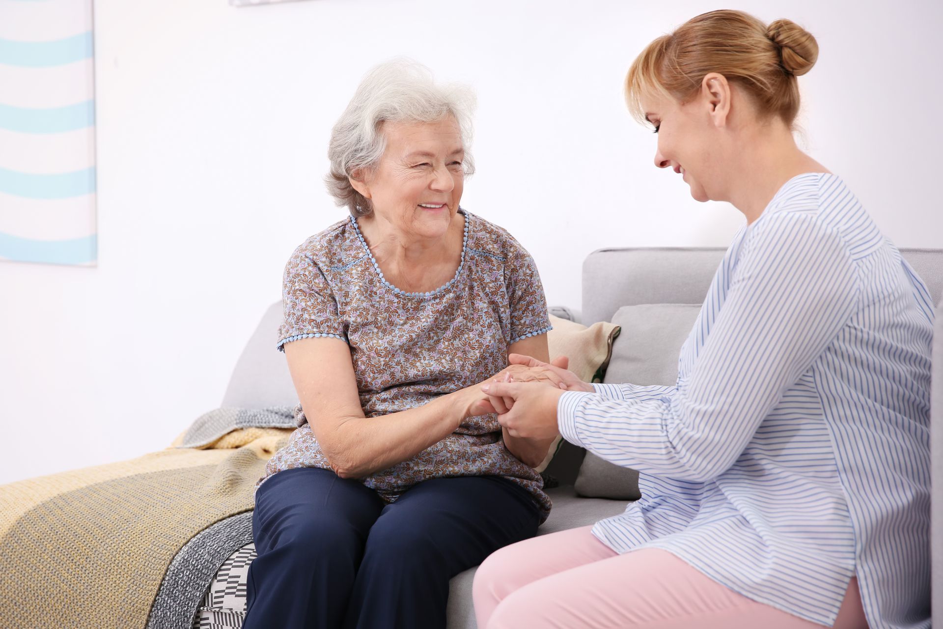 A caregiver is holding hands with a smiling elderly woman on a sofa indoors.