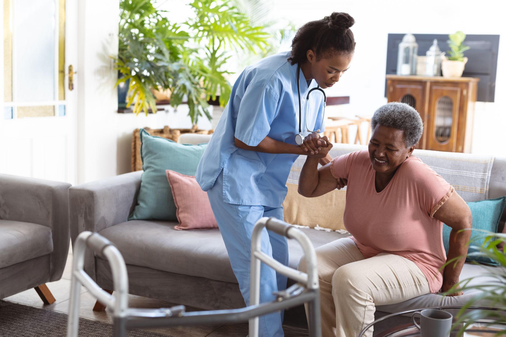 A female nurse helps a female senior standing up from a sofa. A female nurse helps a female senior standing up from a sofa.