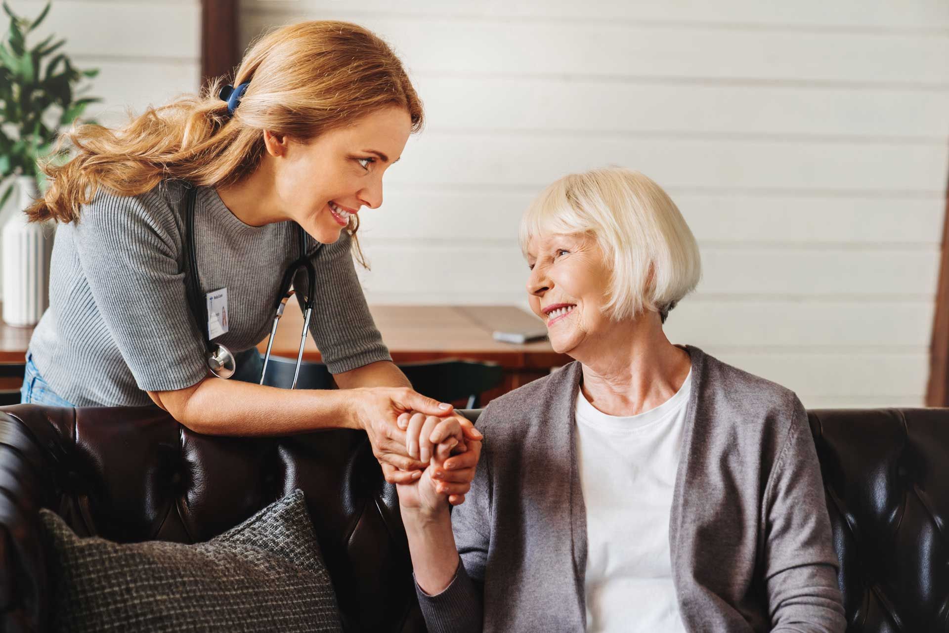 Woman helping an elderly woman sitting on a couch, holding hands, smiling in a living room.