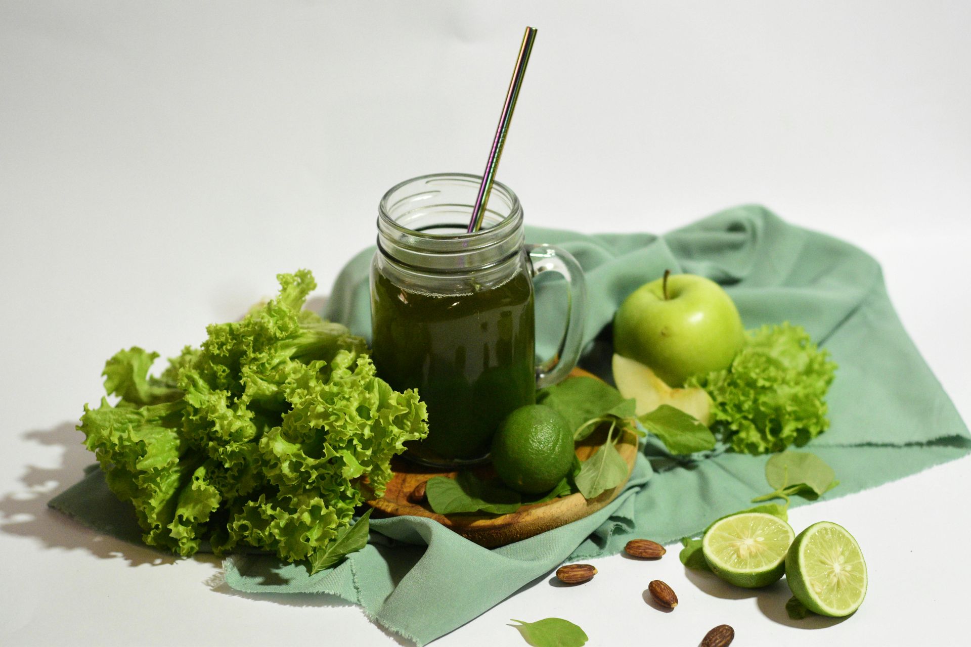 Green juice in a glass jar with straw, surrounded by kale, apple, cucumber, lime, and mint on a white surface.